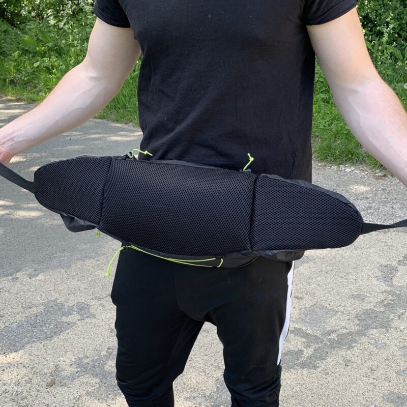 Torso of a person in a black t-shirt holding a wide black padded waist pack with mesh front and neon-green zipper pulls, photographed outdoors on a paved path with grass behind them.