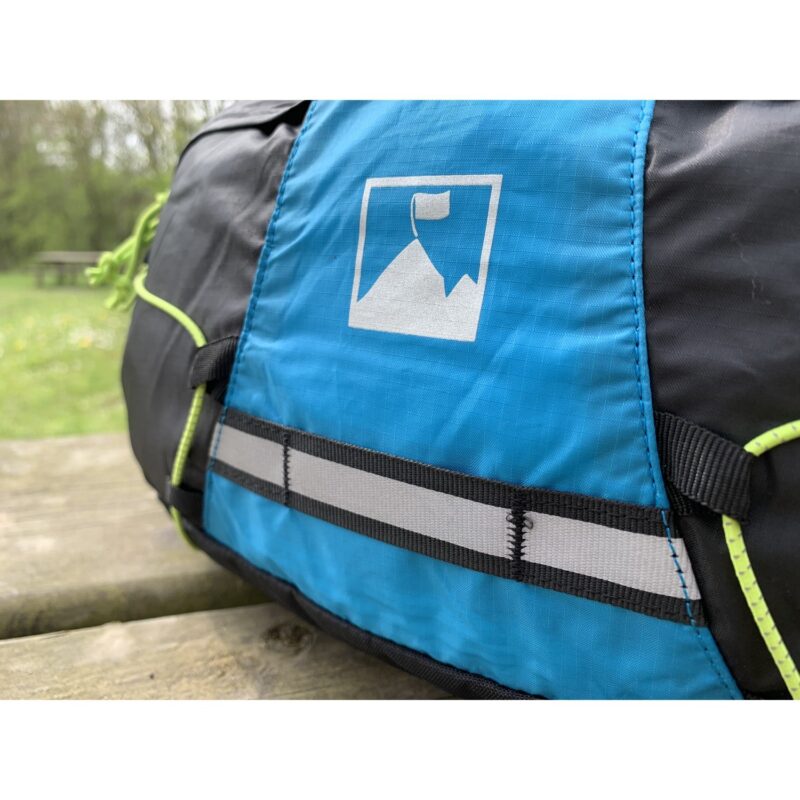 Close-up of a blue and black hiking pack on a wooden picnic table, showing a white mountain-with-flag logo, a reflective horizontal strap and neon green cord with a blurred grassy park background
