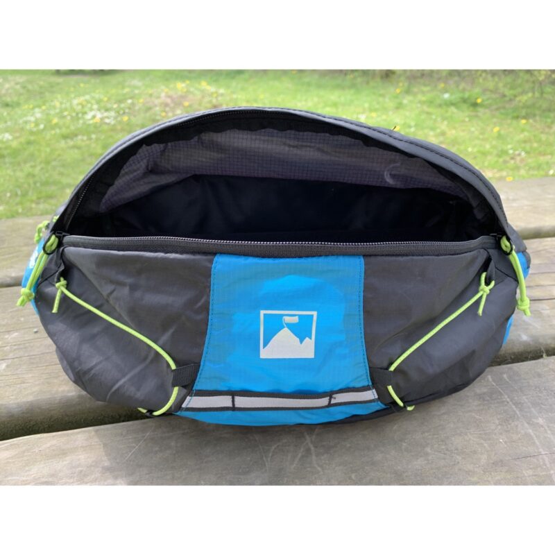 Open black and blue waist pack resting on a wooden picnic bench, showing an empty black interior, a front blue panel with a white mountain-and-flag logo, neon green bungee cord accents and a top zipper, with grass in the background.