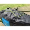 Black and blue backpack resting on a wooden picnic bench, close-up of a green elastic bungee cord laced in an X across the pack's top panel with out-of-focus grass in the background.
