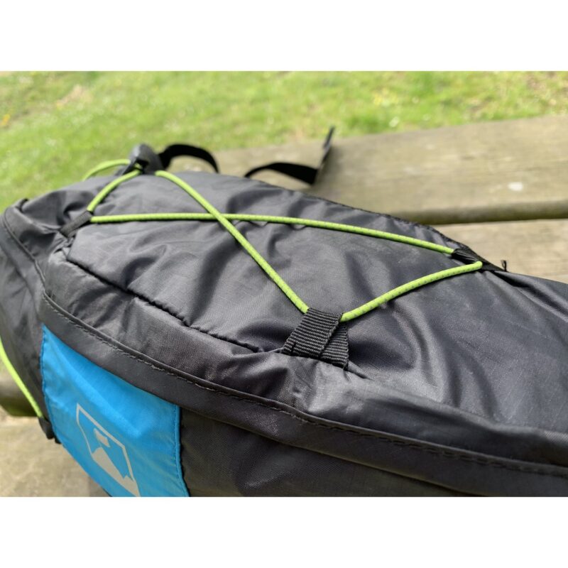 Black and blue backpack resting on a wooden picnic bench, close-up of a green elastic bungee cord laced in an X across the pack's top panel with out-of-focus grass in the background.