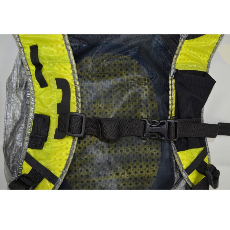 zoom_Laser_Ultra_5_Pack_3-1.jpg Close-up of a backpack harness showing a central black plastic chest buckle and adjustable webbing across a padded mesh back panel with yellow trim, dotted ventilation pattern and silver-edged fabric.