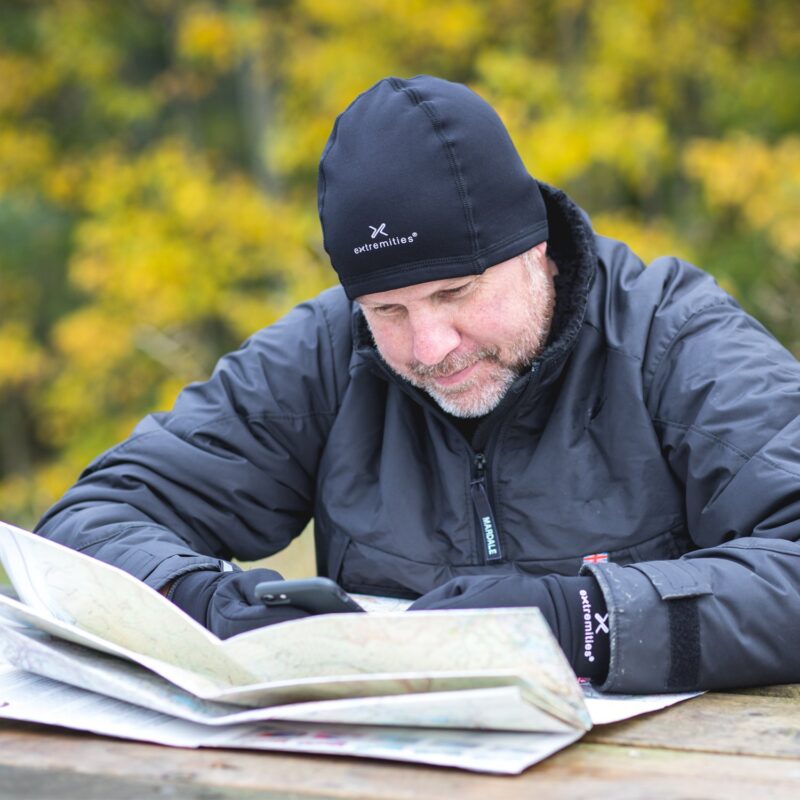zoom_Primaloft_Stretch_Beanie_1-3.jpg Man wearing a black Extremities beanie, gloves and insulated jacket leans over an open map on a wooden picnic table while checking his phone, with yellow autumn trees blurred in the background.