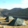 zoom_Quasar_Tent-1.jpg Two green dome tents pitched on a windswept grassy plateau beside a steep layered canyon, a camper sitting on the ground among gear with rolling hills and a bright blue sky in the background.