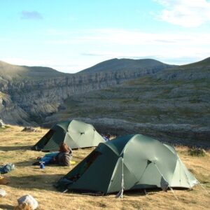 Two green dome tents pitched on a windswept grassy plateau beside a steep layered canyon, a camper sitting on the ground among gear with rolling hills and a bright blue sky in the background.