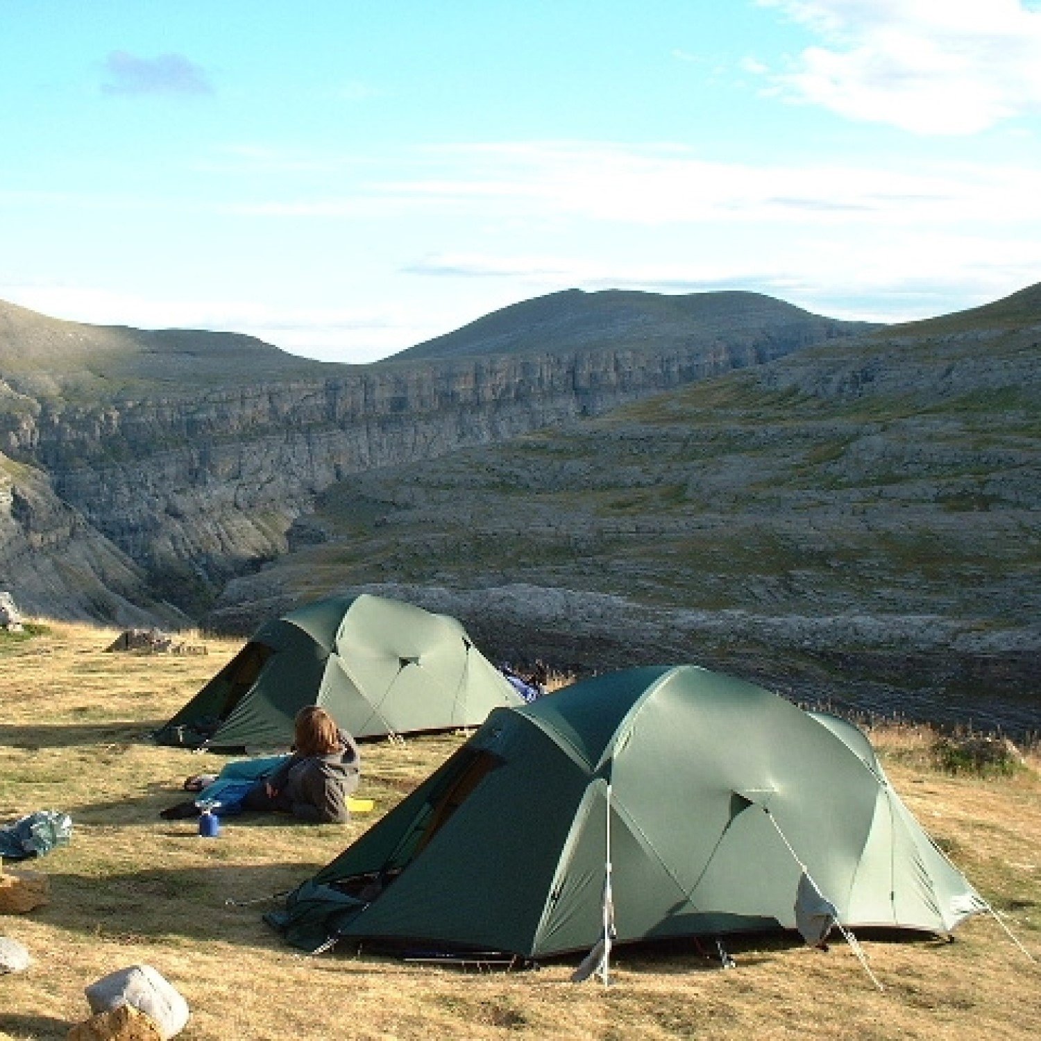 Two green dome tents pitched on a windswept grassy plateau beside a steep layered canyon, a camper sitting on the ground among gear with rolling hills and a bright blue sky in the background.