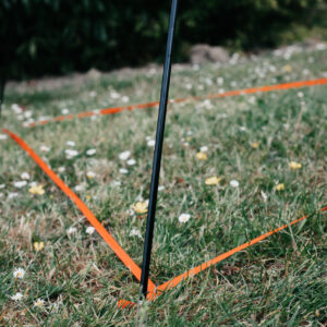 Black stake with orange marking tape on grass, indicating a defined area for landscaping or construction. Wildflowers are scattered throughout the grassy field, adding a natural touch to the scene.