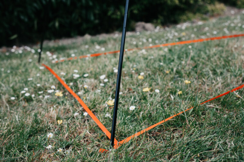 Black stake with orange marking tape on grass, indicating a defined area for landscaping or construction. Wildflowers are scattered throughout the grassy field, adding a natural touch to the scene.