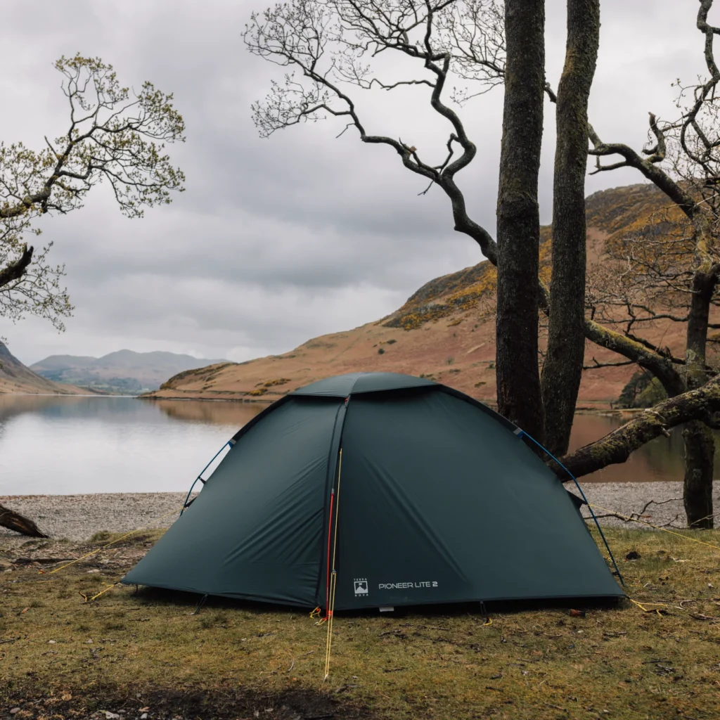 A green dome-style camping tent staked on a rocky, grassy upland at dusk with pale sky and layered blue mountain ridges in the distance.