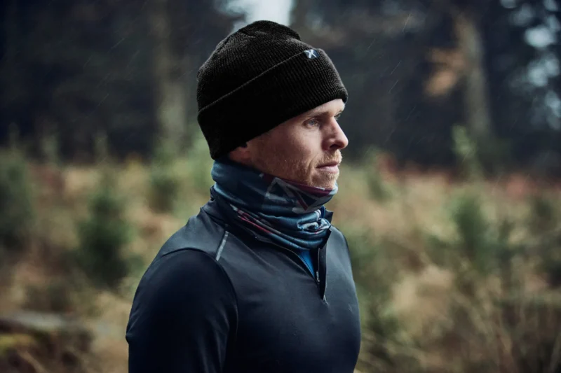 Contour Waterproof Eco Polyana Beanie Lifestyle Man wearing a black beanie and a blue scarf, standing outdoors in a natural setting, with trees in the background, showcasing active outdoor apparel suitable for cold weather.
