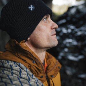 Man wearing a black beanie and an orange jacket, gazing thoughtfully while surrounded by a rocky outdoor setting.