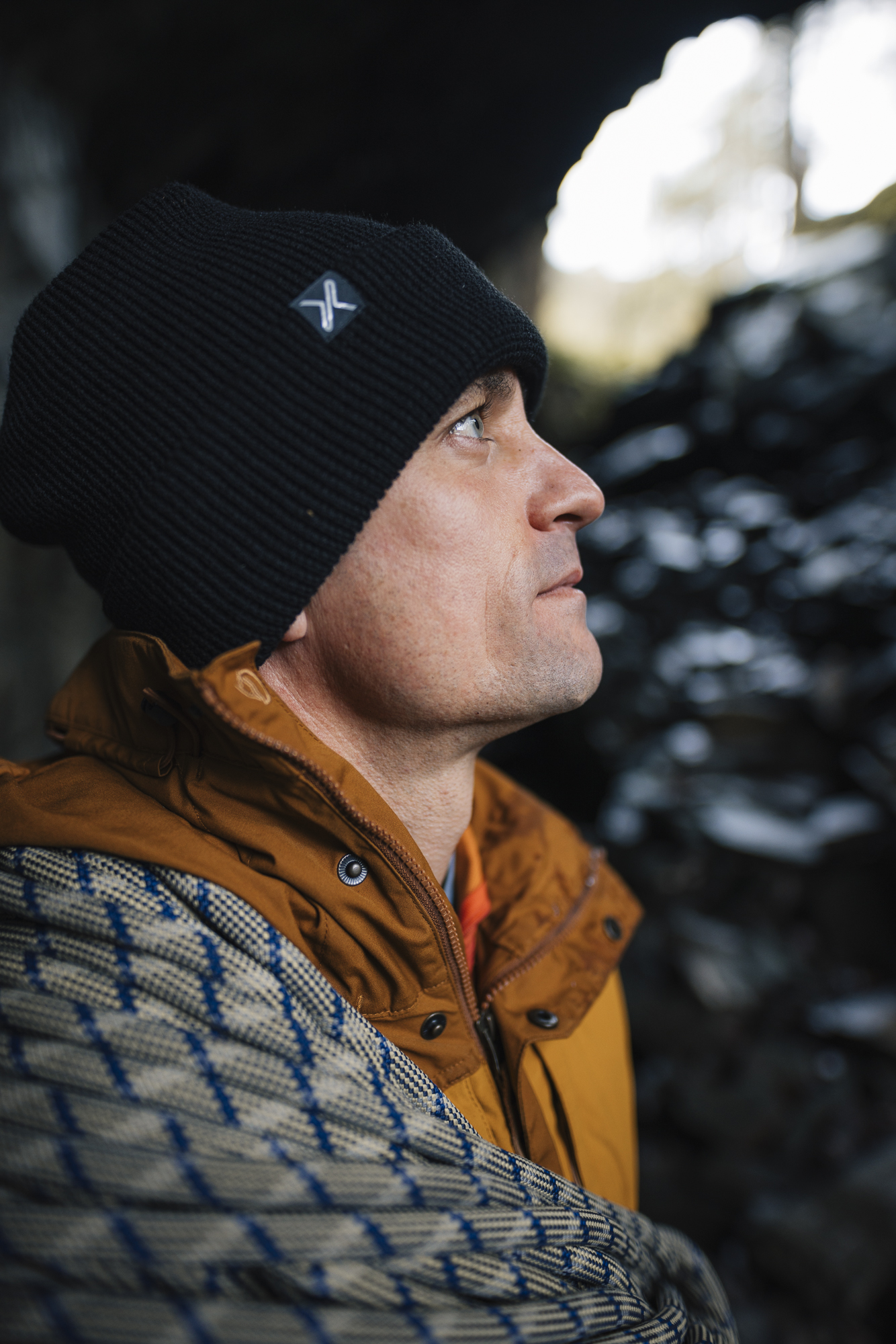 Man wearing a black beanie and an orange jacket, gazing thoughtfully while surrounded by a rocky outdoor setting.
