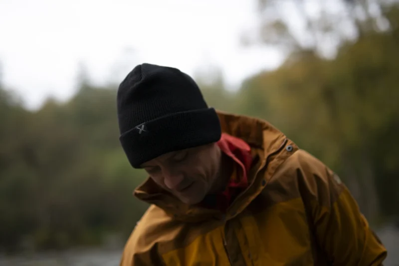 Contour Waterproof Eco Polylana Beanie (7) A person wearing a black beanie and a yellow rain jacket, looking down thoughtfully in an outdoor setting with blurred greenery in the background.