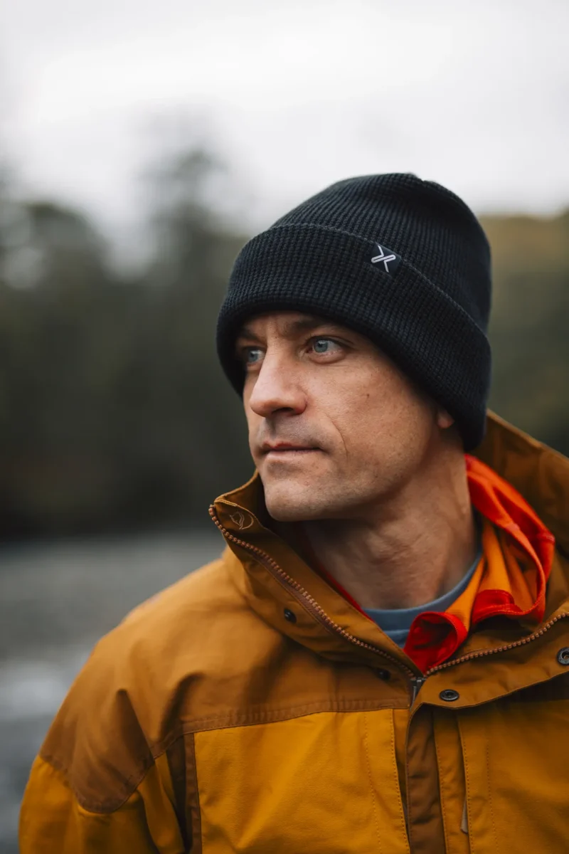 Contour Waterproof Eco Polylana Beanie (8) Man wearing a black beanie and orange jacket, looking thoughtfully into the distance in an outdoor setting.