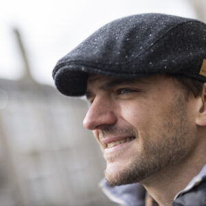Man wearing a stylish flat cap, smiling outdoors with a blurred background.