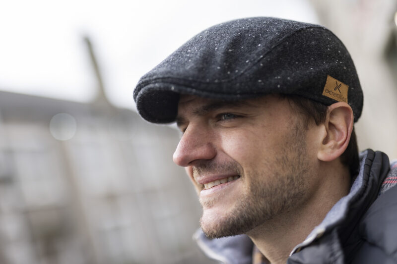 Man wearing a stylish flat cap, smiling outdoors with a blurred background.
