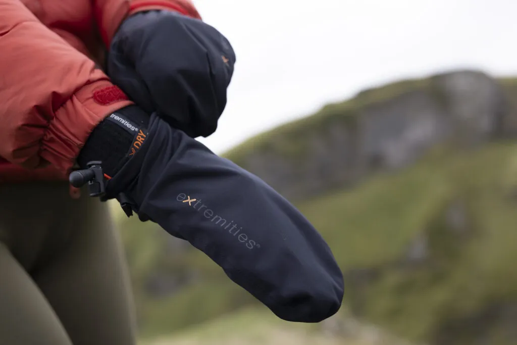 Monsoon Mitt (5) Close-up of a person's hand wearing a waterproof black mitten, designed for outdoor activities in cold weather, with a blurred natural landscape in the background.