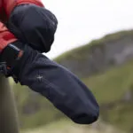Close-up of a person's hand wearing a waterproof black mitten, designed for outdoor activities in cold weather, with a blurred natural landscape in the background.