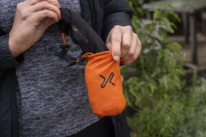Person holding a compact orange pouch with a black drawstring, designed for easy portability, set against a natural outdoor background.