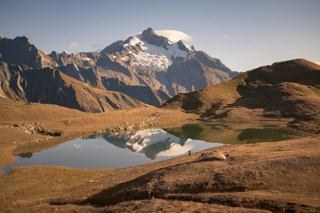 Scenic mountain landscape with a snow-capped peak reflected in a tranquil lake, surrounded by rugged terrain and golden grasses under a clear blue sky.