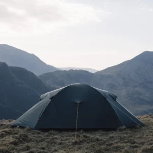 A green dome-style camping tent staked on a rocky, grassy upland at dusk with pale sky and layered blue mountain ridges in the distance.