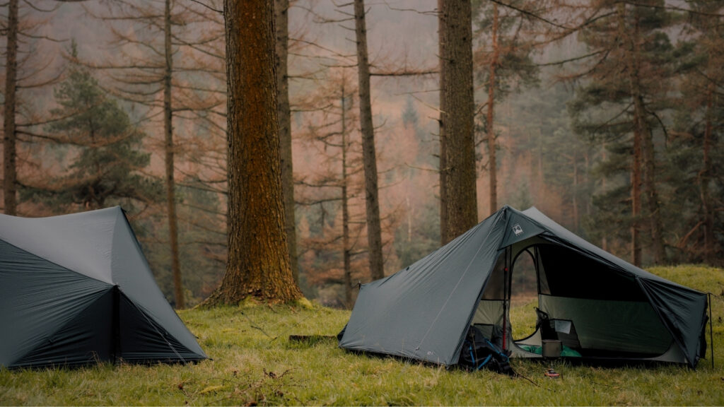 Two Terra Nova iO ultralight trekking pole tents pitched on a grassy forest clearing, showcasing their lightweight shelter design in a woodland setting.