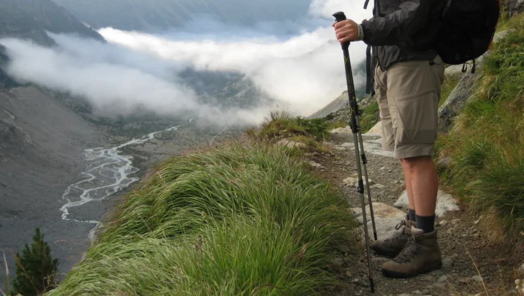 Partial view of a hiker's legs and hands holding a trekking pole on a rocky mountain path, wearing hiking boots, shorts and a jacket, overlooking a cloud-filled valley with a winding river and wind-swept grasses in the foreground