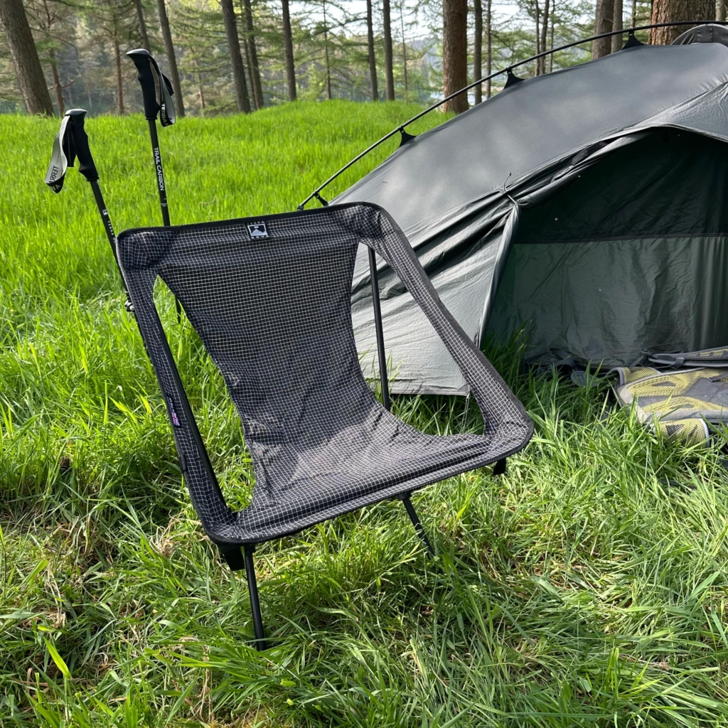 A green dome-style camping tent staked on a rocky, grassy upland at dusk with pale sky and layered blue mountain ridges in the distance.